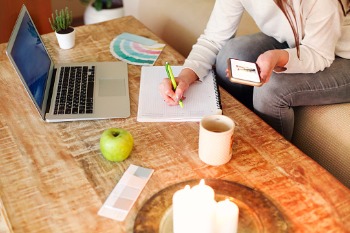 Eine Fernstudentin für Ernährung sitzt im Wohnzimmer, auf dem Tisch stehen ein Laptop, sie macht sich Notizen und hat neben sich einen grünen Apfel