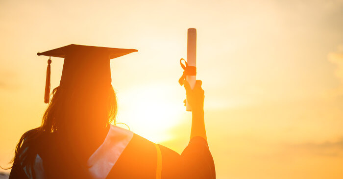 Eine Masterabsolventin steht im warmen Gegenlicht der Sonne und hält ihre Abschlussurkunde hoch